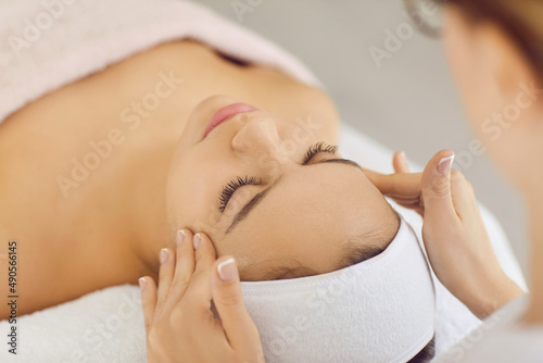 Relaxed woman relieves stress while receiving professional facial and head massage in spa salon. Close up of female masseur hands doing massage to client lying with eyes closed in white bandage.