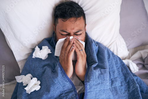 An overhead shot of a sick man blowing his nose while lying in bed