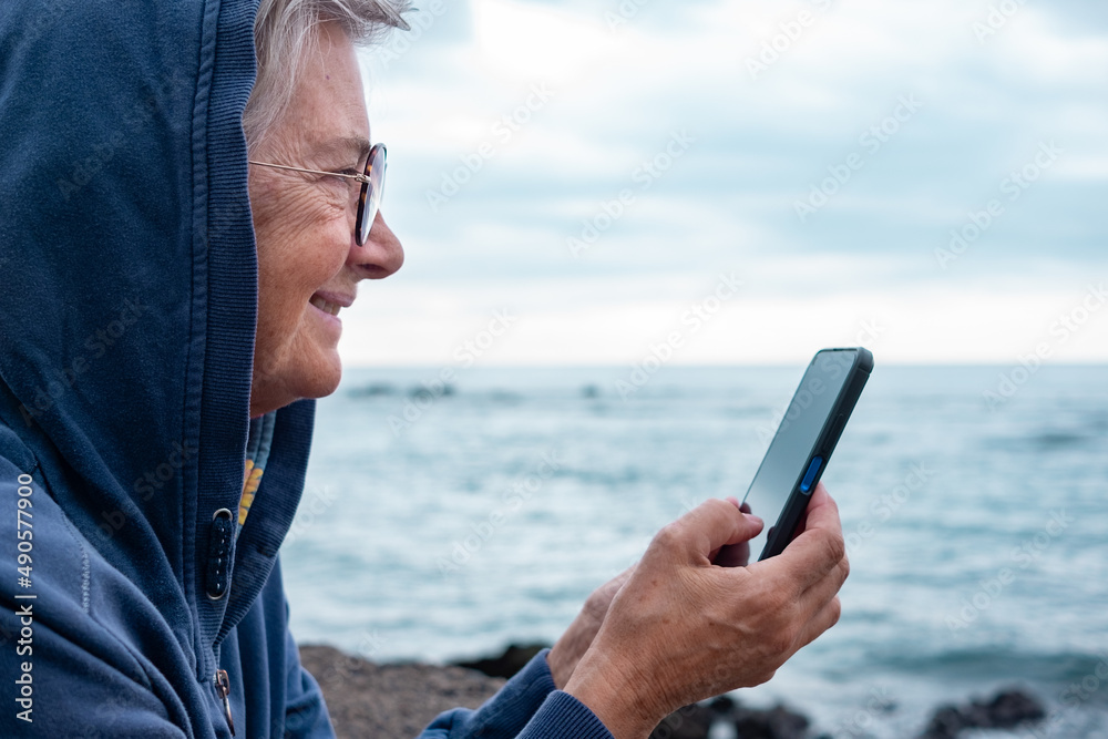 Close up portrait of mature senior woman enjoying retirement at sea. Caucasian elderly female standing on the beach in a cloudy day using mobile phone enjoying tech and social. Horizon over water