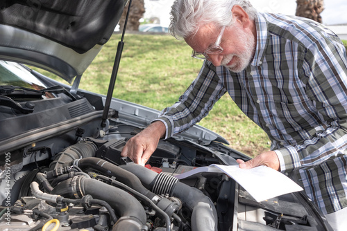 Elderly man driver stopped in the road due to a car breakdown opens the hood and looks at the instruction booklet to understand what is broken. Senior citizen waiting for roadside assistance