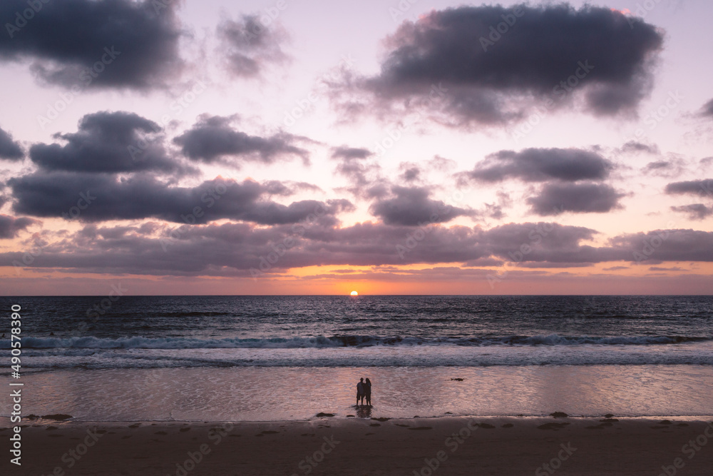 Couple stands on beach at sunset as sun dips below horizon