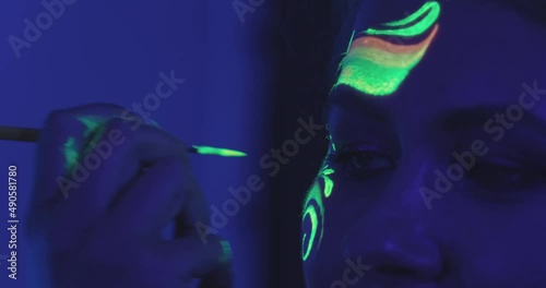 Makeup artist draws on face of young woman with neon paint. Bright colors glowing at ultraviolet illumination of dark club. Thematic party closeup photo