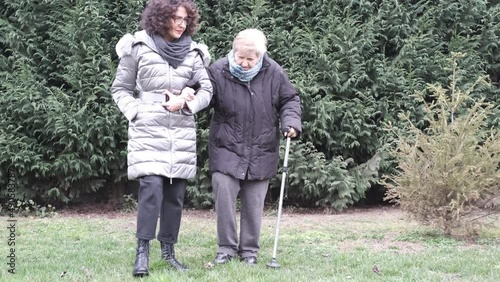 An old woman walks in the park with the help of a caregiver. Smiling caregiver walks and chats with senior woman. Walk around the park with a walking stick in her hand. Old age.

