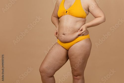 Cropped image of overweight woman, tucking, hiding fat naked big excessive belly with navel in yellow pants. Dangling down stomach, big size tummy. Drag away of abdomen. Go on diet, liposuction photo