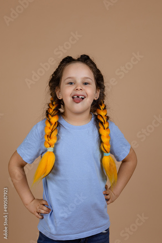Mischievous little shining girl with slight smile showing tongue looking at camera misbehaving having kanekalon braids on beige background wearing blue t-shirt. Bad habits.