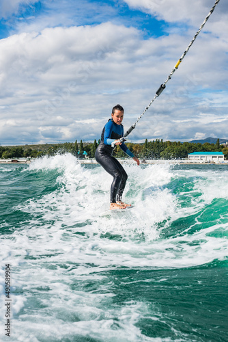Female in wetsuit riding the waves using of tow rope behind a boat on sunny day. Watersport concept. Young athletic woman learning wakesurfing and perfecting tricks © tgordievskaya