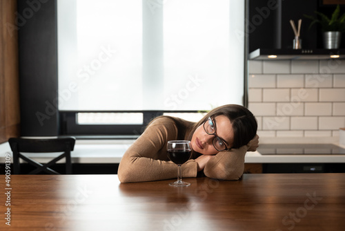 Young sad depressed woman sitting alone at home drinking wine feeling abandoned. Anxious female suffering from depression after relationship breakup drinking alcohol drink in kitchen try not to cry.