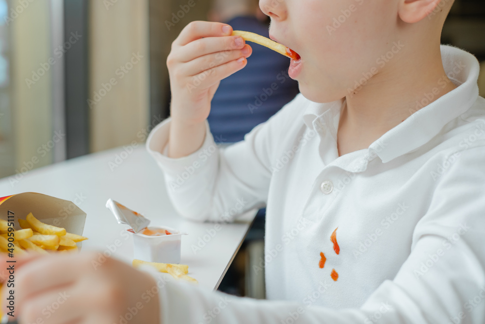 Close up of dirty ketchup stains on white kid clothes. Boy eating french fries potato chips and tomato sauce at the table in a fast food restaurant. Kids eat unhealthy fat food. Close up of dirty ketchup stains on white kid clothes. Boy eating french fries potato chips and tomato sauce at the table in a fast food restaurant. Kids eat unhealthy fat food.