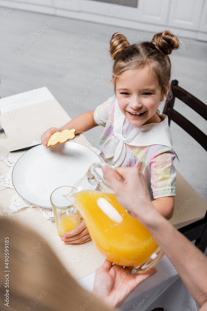 blurred woman pouring orange juice near pleased daughter holding easter cookie. blurred woman pouring orange juice near pleased daughter holding easter cookie.