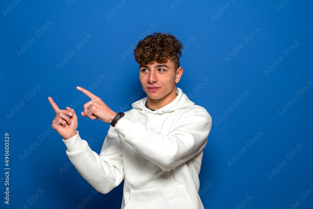 Smiling young man 20s in casual white sweatshirt posing isolated on blue wall background studio portrait. People candid emotions lifestyle concept. Mock up copy space. Pointing fingers to the side. Smiling young man 20s in casual white sweatshirt posing isolated on blue wall background studio portrait. People candid emotions lifestyle concept. Mock up copy space. Pointing fingers to the side.