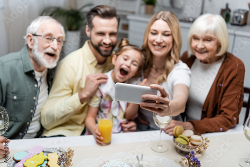 woman taking selfie on smartphone with blurred family during easter dinner.