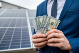 boss in a jacket with hands holding a large sum of dollars on a background of solar panels