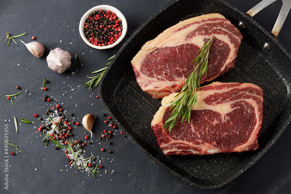 Raw meat, two beef steaks in a grill pan with rosemary and spices on a black background, top view Raw meat, two beef steaks in a grill pan with rosemary and spices on a black background, top view