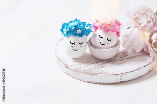 Two eggs with cute smiles and flowers on a wooden stand. Creative eggs on a white background with lights. Beautiful postcard for the Easter holiday photo