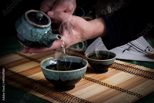 Pouring hot tea from a teapot into a glass on the mat covered table on a dark wooden background.