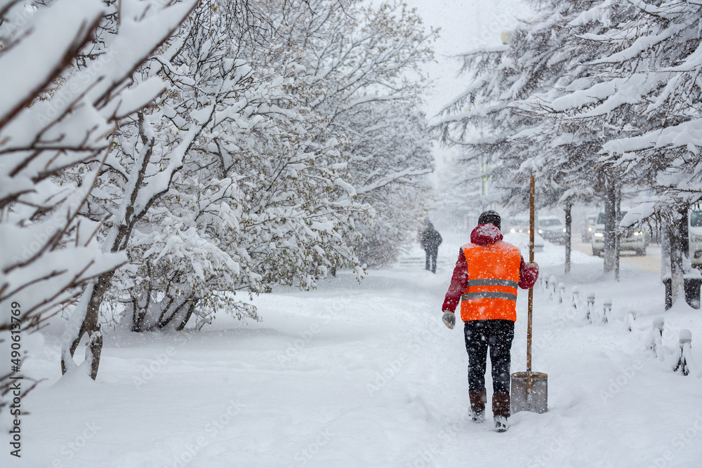 A janitor with a shovel walks along a snow-covered sidewalk. Heavy snowfall in the city. View of the snowy street. Lots of snow on the ground and trees. Cold winter weather. Daily life in the city.