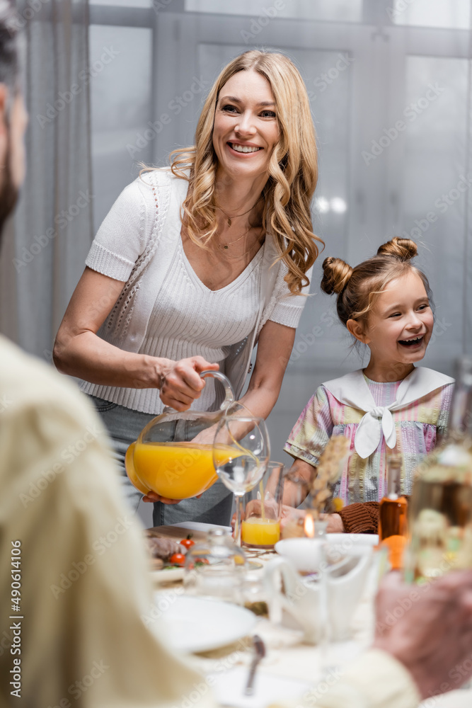 smiling woman pouring orange juice near excited daughter and blurred husband. smiling woman pouring orange juice near excited daughter and blurred husband.