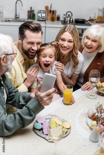 senior man holding smartphone near excited girl with open mouth and happy family.