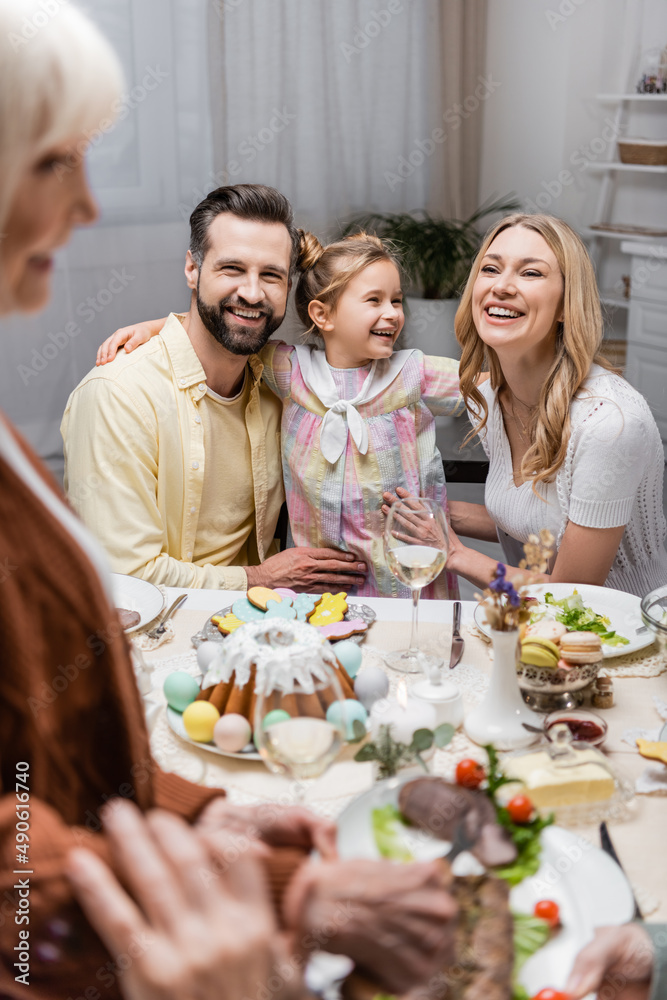 girl smiling near happy parents during easter dinner on blurred foreground.