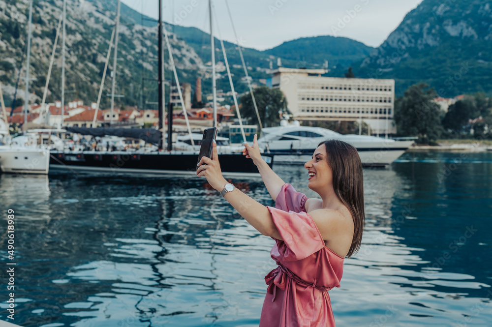 Woman in a pink silky dress taking selfie with a smartphone while walking in a marina Woman in a pink silky dress taking selfie with a smartphone while walking in a marina