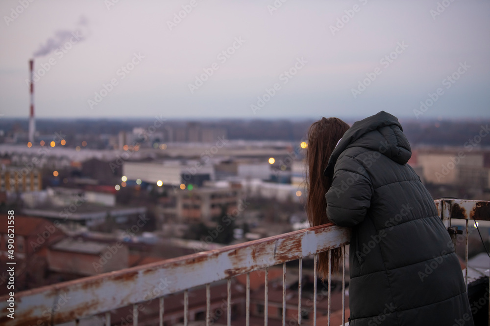 Depressed sad young female in jacket with hoodie standing on the edge of the fence on residential building rooftop. Suicide and major depressive disorder concept. Contemplating suicide.