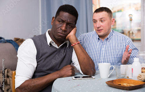 Two sad interracial men sitting at table after conflict at home interior