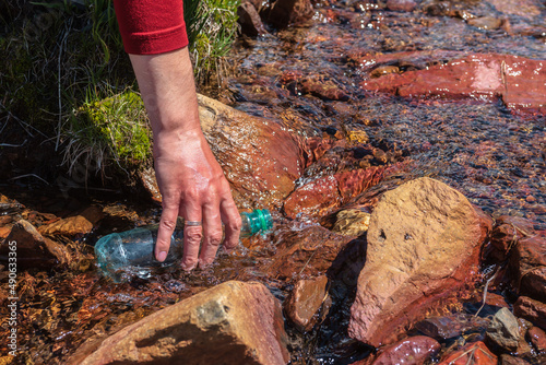 Man replenishes water from mountain stream in bright sun. Tourist fills bottle with mineral water in sunny day. Hand with bottle in clear water stream in sunlight. Replenishment of water during hike.