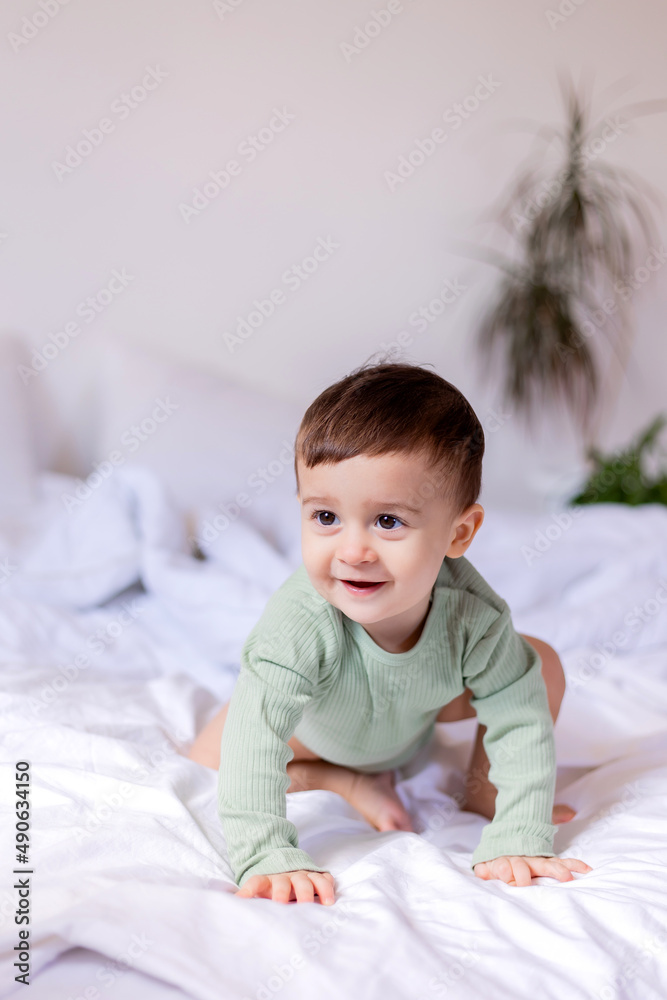 cheerful baby in a green cotton bodysuit is lying on his stomach on a white bed cheerful baby in a green cotton bodysuit is lying on his stomach on a white bed