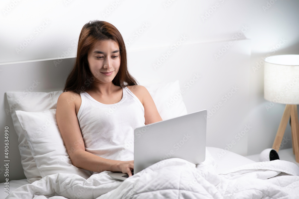 Beautiful Asian woman in pajamas using laptop sitting on the bed in the house. Beautiful Asian woman in pajamas using laptop sitting on the bed in the house.