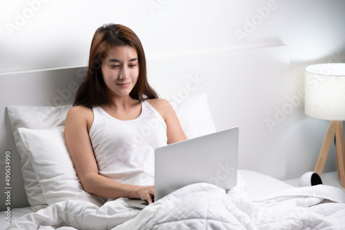 Beautiful Asian woman in pajamas using laptop sitting on the bed in the house.