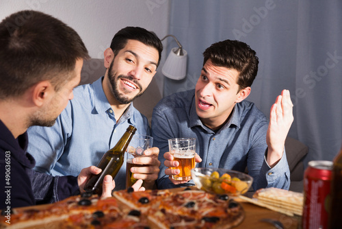 Three adult cheerful men talking and laughing while enjoying beer and pizza at home