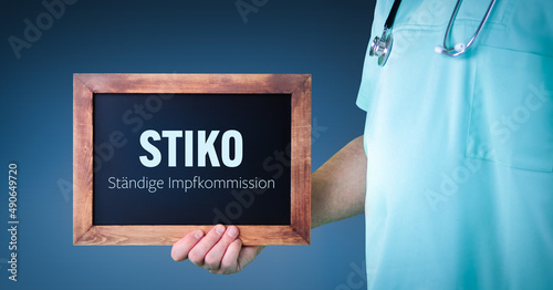 STIKO (Ständige Impfkommission). Arzt zeigt Schild/Tafel mit Holz Rahmen. Hintergrund blau