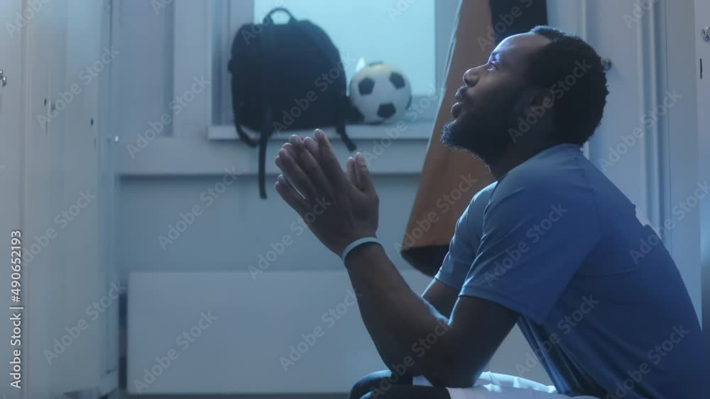 Side view tilting up of young Black male athlete sitting by lockers in dressing room, getting mentally ready, being nervous before match early in morning