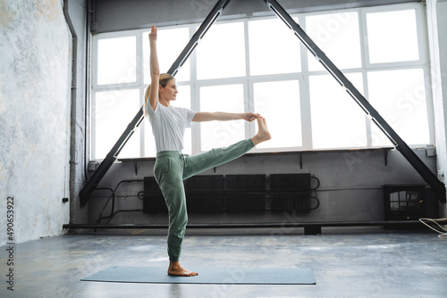 A sporty girl stands on a yoga mat. Girl performs stretching and assanas. Complex poses from yoga. Bright yoga studio with wide windows