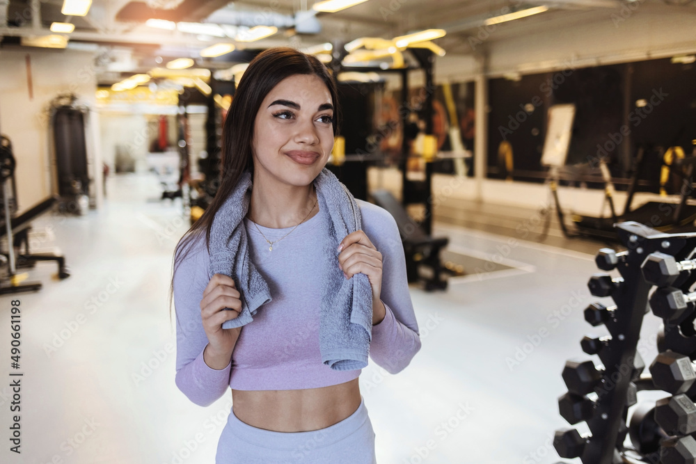 Fit young woman with a towel at health club. Female relaxing after workout. Portrait of charming smiling young fitness girl with a towel and posing while looking at the camera in the gym.