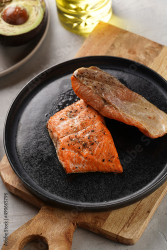 Two grilled fish steaks - red salmon on black round plate on wooden board, olive oil, avocado on grey background