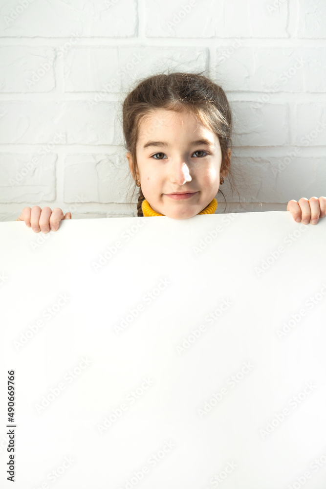 A sad girl holds a white sheet of paper - mock-up for advertising, slogan, inscription. Copy space is in children's hands, the child is upset and hid. A sad girl holds a white sheet of paper - mock-up for advertising, slogan, inscription. Copy space is in children's hands, the child is upset and hid.