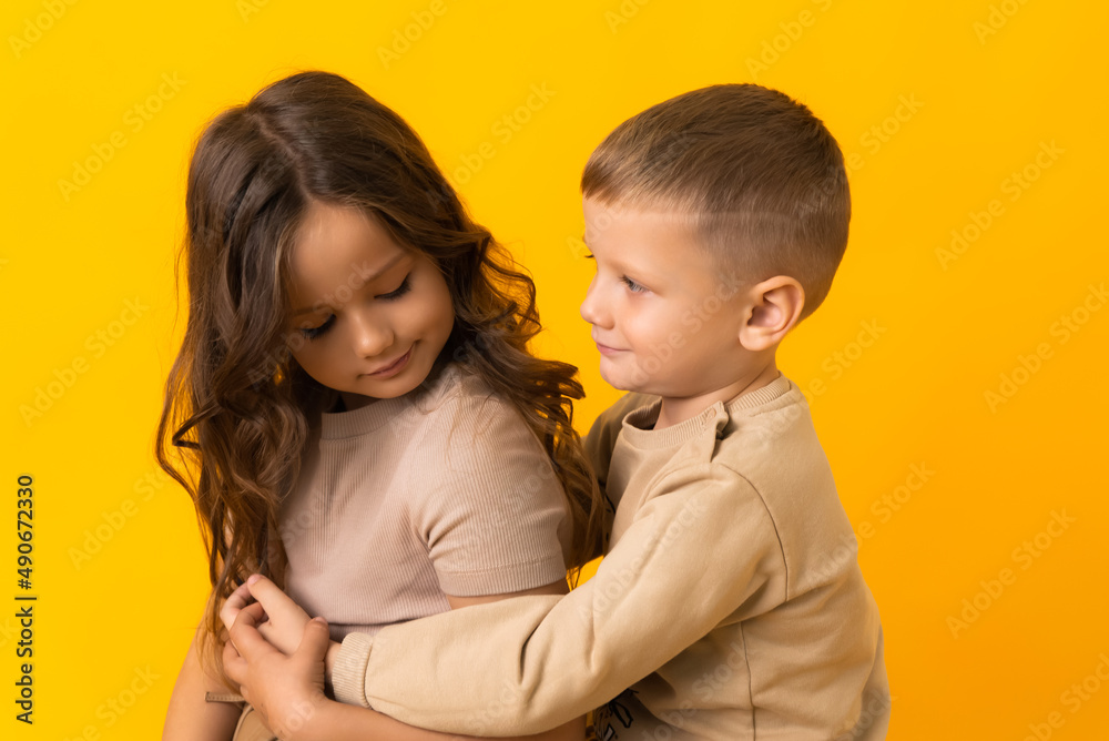 A little brother is hugging his older sister in the studio on a yellow background A little brother is hugging his older sister in the studio on a yellow background