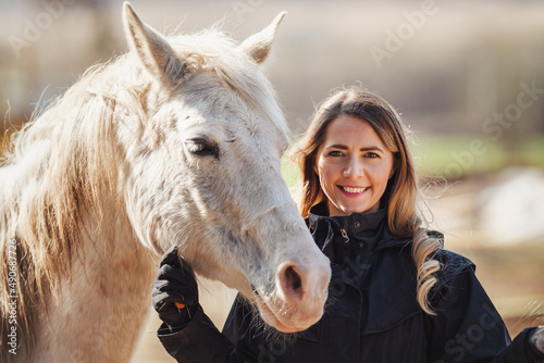 Young woman in black riding jacket standing near white Arabian horse smiling happy, closeup detail © Lubo Ivanko