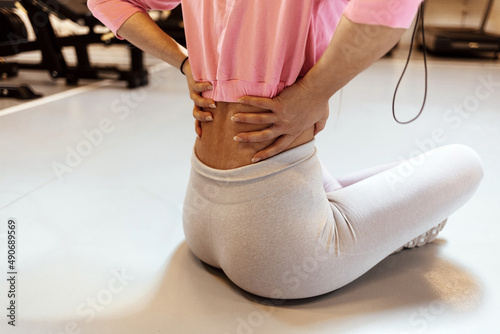 Shot of a sportswoman with a lower back injury. Young woman resting with back pain, sitting on the floor at a fitness center. Female hand touching body part with pain from wrong action workout in gym.