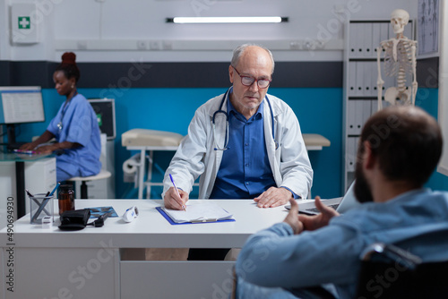 Young man with disability having medical consultation with doctor for medical advice and support. Patient sitting in wheelchair while discussing with specialist about healthcare.