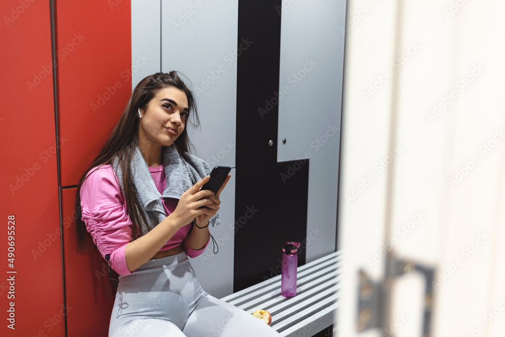Close-up of Caucasian woman using smart phone and text messaging in dressing room at the gym. Happy female athlete listening music and texting on cell phone in dressing room at the gym.