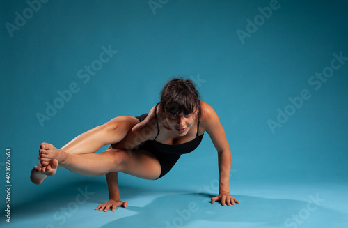 Personal trainer in sportwear working at arm balance practicing yoga position in studio with blue background. Active fit woman stretching body muscles during sport workout