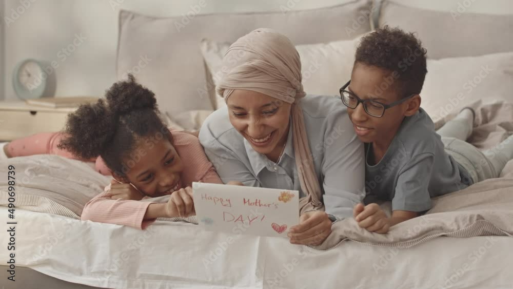 Slowmo of smiling Biracial woman getting handmade postcard on Mothers day from her two 8 year old kids, lying on bed together in morning Slowmo of smiling Biracial woman getting handmade postcard on Mothers day from her two 8 year old kids, lying on bed together in morning