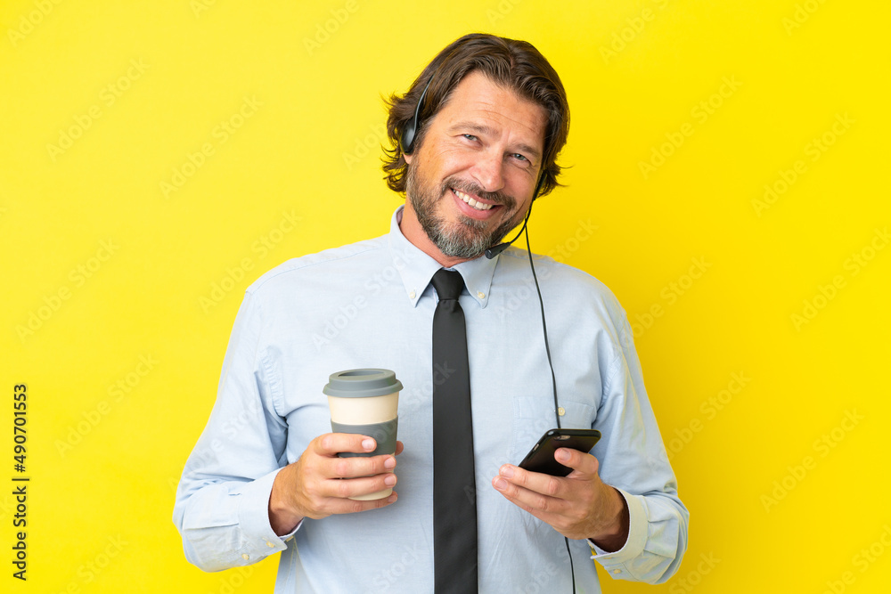 Telemarketer dutch man working with a headset isolated on yellow background holding coffee to take away and a mobile Telemarketer dutch man working with a headset isolated on yellow background holding coffee to take away and a mobile