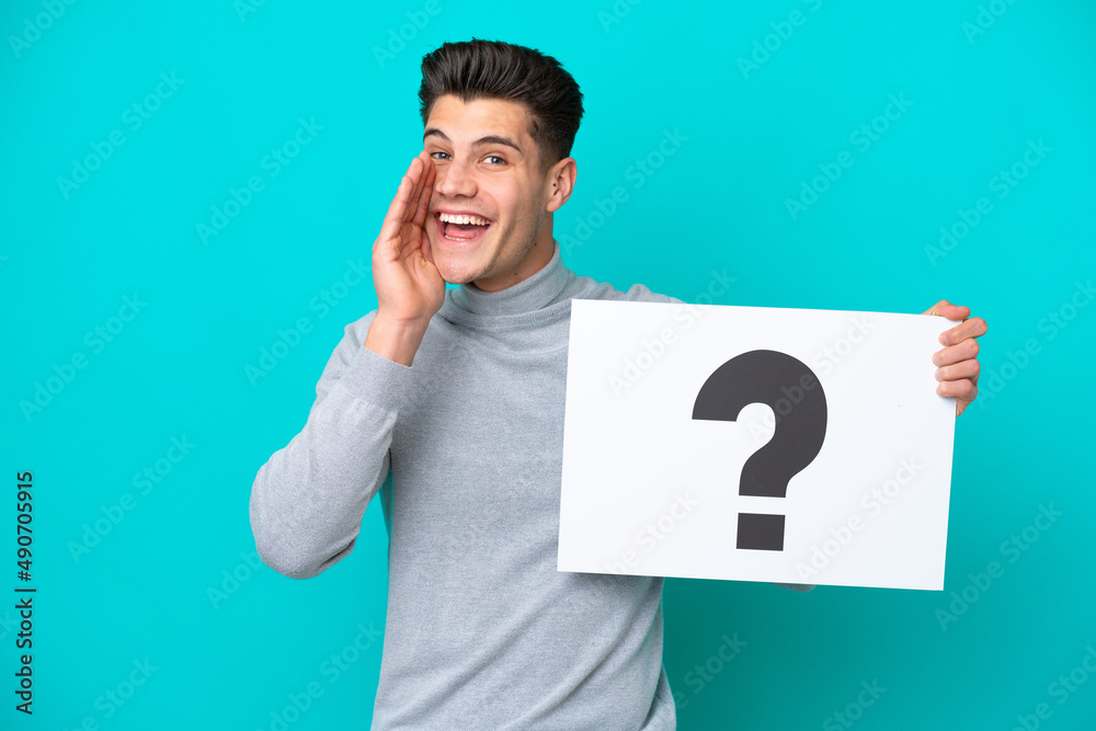 Young handsome caucasian man isolated on blue bakcground holding a placard with question mark symbol and shouting