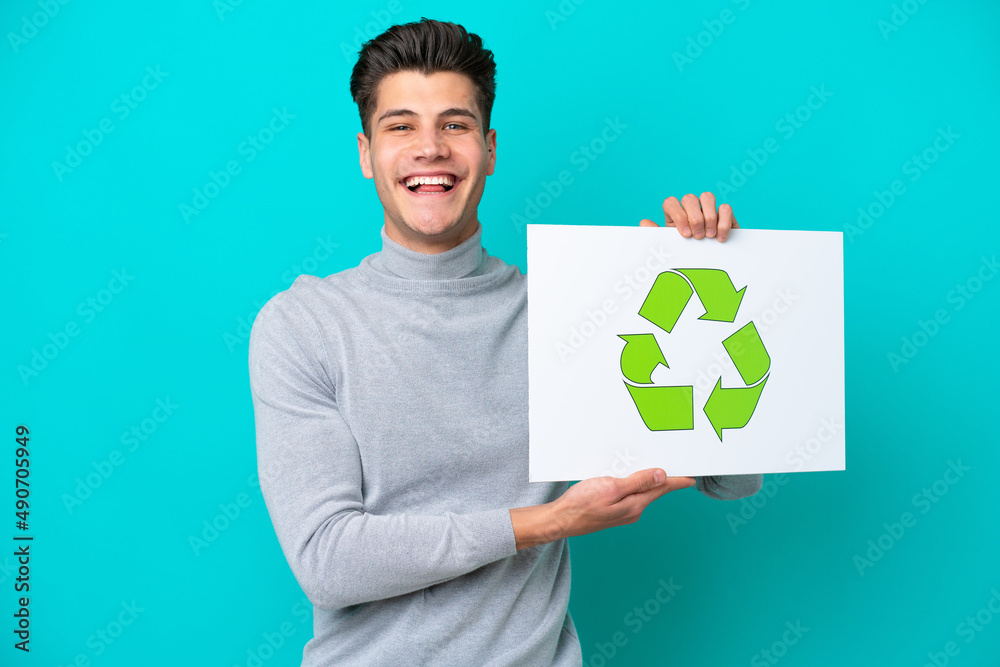 Young handsome caucasian man isolated on blue bakcground holding a placard with recycle icon with happy expression
