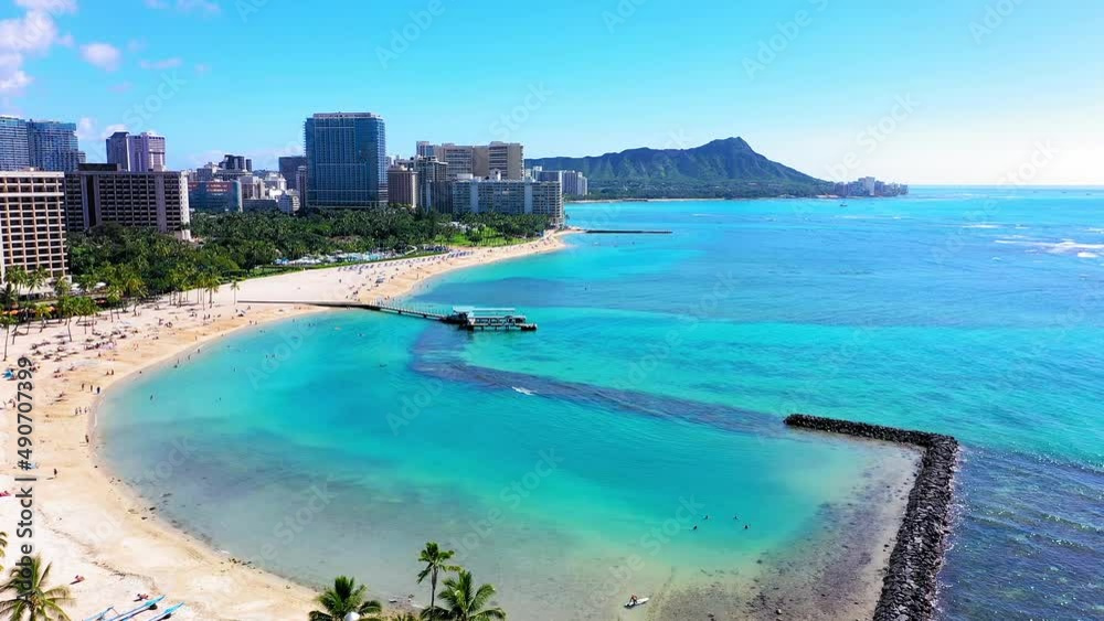 Aerial drone view flying down the beach and towards the Diamond Head Volcano with a great view of hotels, Pacific Ocean, and lots of palm trees. Perfectly clear water with a nice view of the reef.