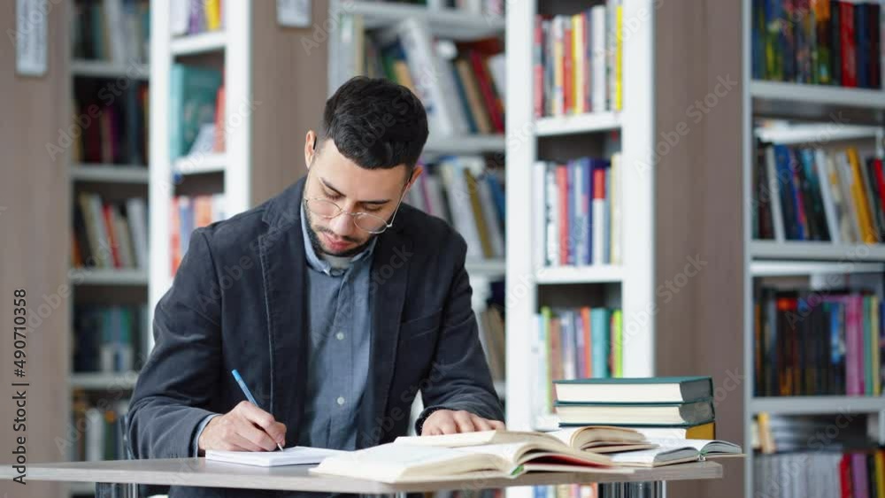 Student wearing eyeglasses writing homework in library, speaking aloud his ideas, heap of books lying beside, bookcases on blurred background. Concept of study Student wearing eyeglasses writing homework in library, speaking aloud his ideas, heap of books lying beside, bookcases on blurred background. Concept of study