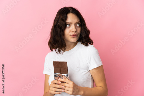 Teenager Ukrainian girl isolated on pink background taking a chocolate tablet and having doubts